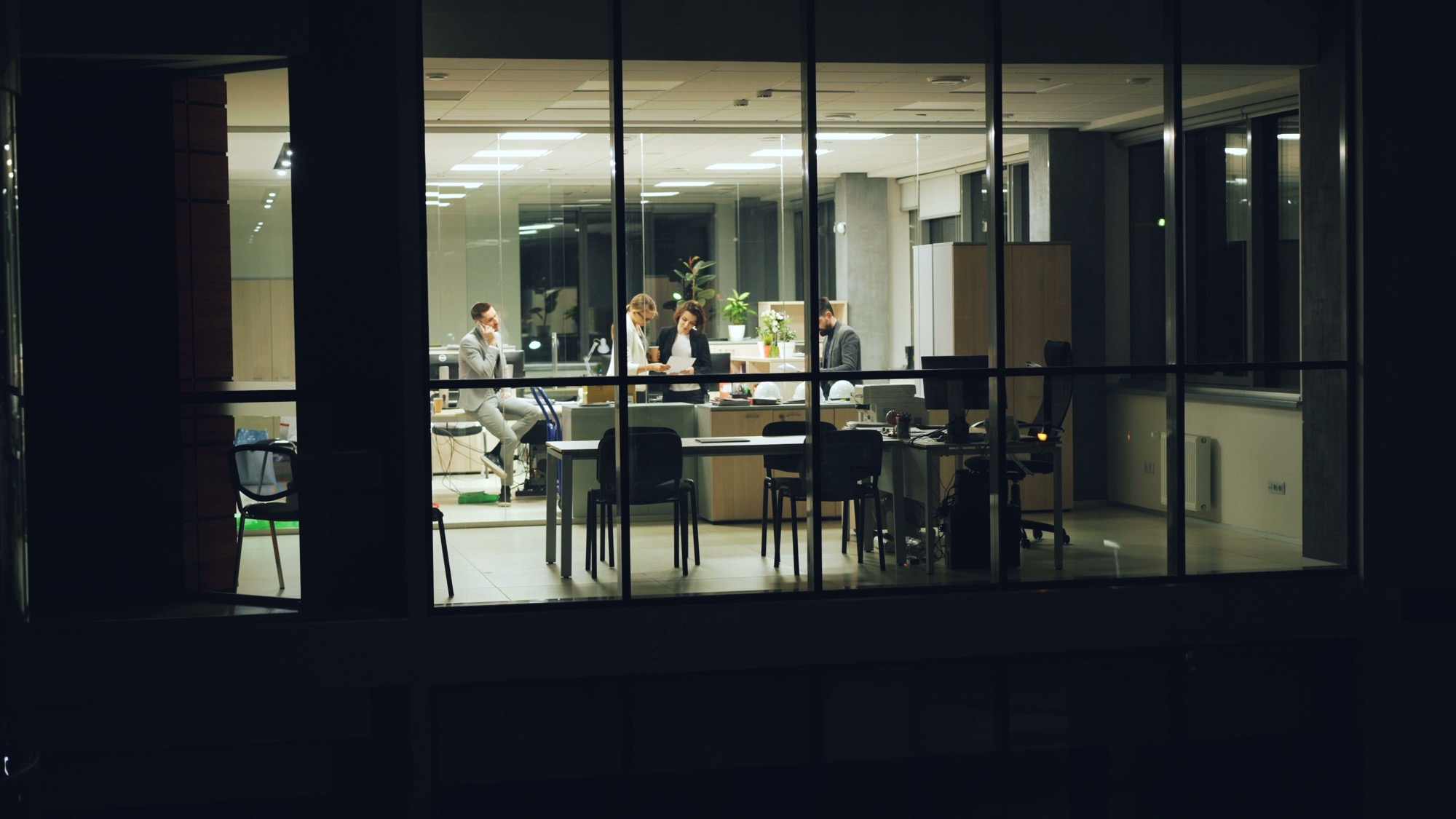 Lit office seen through glass after dark, people still at the table